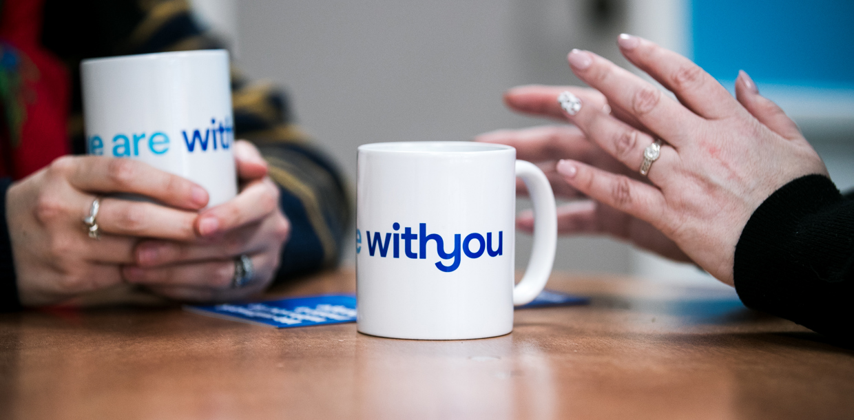 Two people sitting at a table, holding mugs with the text "WithYou" visible on one of them.